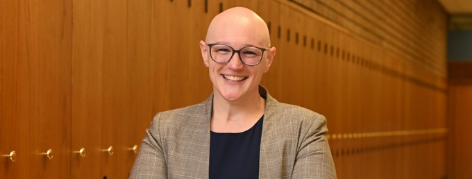 woman wearing glasses, smiling, standing in a hallway with lockers. 