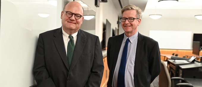 two men standing in a classroom next to a white board. 