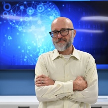Man wearing glases, smiling, standing in front of a large screen mounted on a wall. 