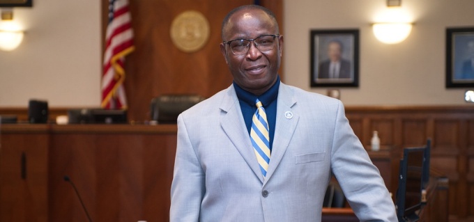Man standing in a courtroom, wearing a blue suit. 