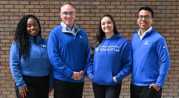 Four individuals standing in front of a brick wall, all wearing University at Buffalo School of Law apparel in shades of blue. Two are in quarter-zip pullovers with UB branding, one in a hoodie with &ldquo;University at Buffalo School of Law&rdquo; printed on the front, and one in a crewneck sweatshirt with the UB logo. They are posed side by side, facing forward. 