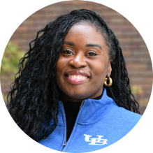 Close-up of a person wearing a royal blue University at Buffalo zip-up jacket with the white &ldquo;UB&rdquo; logo on the left side. The jacket is partially zipped, revealing a black shirt underneath. The person has long, curly black hair and is wearing gold hoop earrings. The background is softly blurred with earthy tones. 