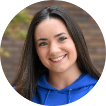 Close-up of a person wearing a bright blue hoodie with the words &ldquo;University at Buffalo School of Law&rdquo; printed in white on the front. The hoodie has drawstrings visible near the neckline. The background shows a brick wall with some greenery and autumn-colored leaves. 