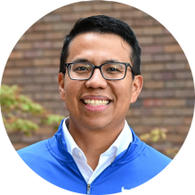 Close-up of a person wearing a bright blue University at Buffalo zip-up jacket over a white collared shirt. The jacket has a small white logo on the left side. The background shows a brick wall with some greenery and autumn-colored leaves. 