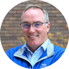 Close-up of a person wearing a blue University at Buffalo jacket with a light gray collar and a white button-down shirt underneath. The jacket features the white &ldquo;UB&rdquo; logo and the words &ldquo;University at Buffalo&rdquo; on the left side. The background shows a brick wall with some greenery and autumn-colored leaves. 