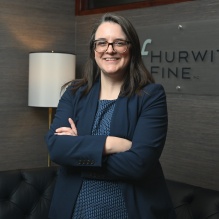 Person standing with arms crossed in a modern office setting. The individual is wearing a navy blazer over a patterned blue top. Behind them is a wall with a mounted sign that reads &ldquo;Hurwitz Fine P.C.&rdquo; and a floor lamp next to a black tufted leather sofa. 