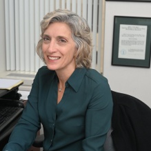 Person seated at a desk in an office, wearing a dark green button-up shirt. The desk has an open planner with handwritten notes, a pair of eyeglasses, and papers. Behind the desk are framed certificates on the wall and vertical blinds covering a window. 