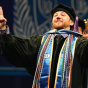 Graduate wearing a black cap and gown adorned with multiple colorful honor cords and stoles raises both hands in a celebratory gesture during a commencement ceremony. A large blue backdrop behind shows the University at Buffalo School of Law emblem featuring a buffalo and shield design. 
