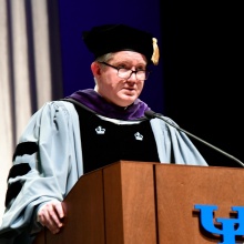 Person wearing academic regalia with a light gray robe and black velvet panels, standing at a wooden podium with a microphone. The podium displays the blue UB logo, and a tall curtain hangs in the background. 