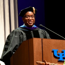 Person in academic regalia standing at a wooden podium with a microphone. The podium displays the blue UB logo and has a ceremonial mace resting on it. A tall curtain hangs in the background, and other individuals in academic attire are partially visible behind the podium. 