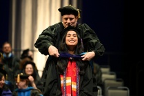 A person in academic regalia places a graduation hood over another individual wearing a red stole with colorful patterns during a commencement ceremony. Several other people in similar regalia are seated in the background, and the setting includes chairs and tall curtains on a stage. 