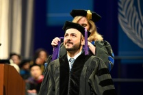 A person in academic regalia is being hooded during a graduation ceremony. Another individual, also in academic regalia, places a purple graduation hood over the person&rsquo;s shoulders. The setting includes a stage with a podium on the left, several people seated in the background, and a large blue banner featuring a laurel design. 