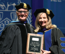 Two individuals in academic regalia stand on stage during a commencement or award ceremony. One person is holding a wooden plaque with engraved text. The background features a large blue banner with an emblem and decorative elements. 
