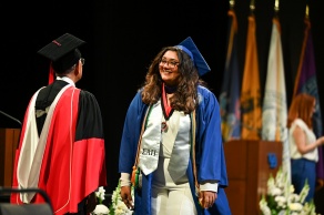 A graduate wearing a blue cap and gown with a white stole and multiple cords stands on stage during a commencement ceremony. The stole has Greek letters and a medallion hangs around the graduate’s neck. A faculty member in black and red academic regalia faces the graduate. In the background, several colorful flags are displayed, along with a podium and floral arrangements. 