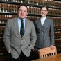 Two people wearing business attire stand in front of shelves filled with rows of legal books in a law library. A wooden table with chairs is visible in the foreground. 