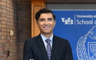 Person wearing a dark suit with a patterned tie stands with arms crossed in front of a wooden reception desk. Behind the desk is a blue banner displaying the University at Buffalo School of Law logo, which includes a buffalo illustration and shield emblem. 