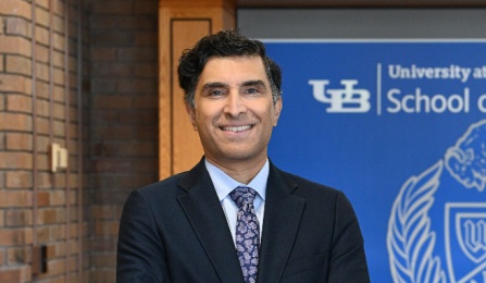 Person wearing a dark suit with a patterned tie stands with arms crossed in front of a wooden reception desk. Behind the desk is a blue banner displaying the University at Buffalo School of Law logo, which includes a buffalo illustration and shield emblem. 