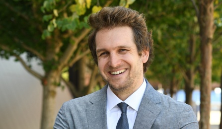 Person wearing a light gray suit with a white dress shirt and dark tie stands outdoors with arms crossed. The background shows green trees, shrubs, and part of a building wall. 