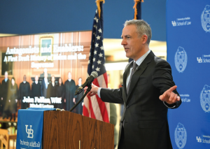 Person in a dark suit and striped tie speaking at a podium with a UB School of Law logo. Behind the podium is an American flag and a large screen displaying the title &lsquo;Antagonists and Enablers: A First Look at Law and Politics&rsquo; with a photo of judges. A blue backdrop with UB School of Law branding is on the right. 