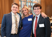 Three people standing together at an indoor event in front of large windows with city buildings visible outside. One person is wearing a blue suit with a striped yellow and blue tie, another is in a royal blue dress, and the third is in a dark suit with a white shirt and red patterned tie. All have visible name tags. 
