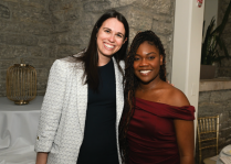 Two people standing indoors in front of a stone wall and a white column. One person is wearing a light textured blazer over a dark outfit, and the other is wearing an off-the-shoulder burgundy dress. A gold decorative lantern and a gold chair are visible in the background. 