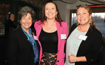 Three people standing together at an indoor event. One person is wearing a black blazer over a blue patterned blouse, another is in a bright pink blazer over a black top with a patterned skirt, and the third is in a black blazer over a light blouse with small dark dots. All have visible name tags. 