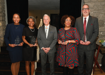Five people standing indoors on steps in front of a black curtain and stone wall. They are dressed in formal attire, including suits and dresses, and each person is holding a glass award. 
