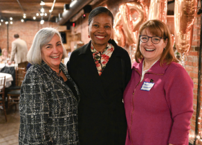 Three people standing close together in a warmly lit indoor space with exposed brick walls and metallic balloon decorations in the background. One person is wearing a patterned jacket, another is in a black outfit with a floral top underneath, and the third is in a bright pink zip-up jacket with a visible name tag. 
