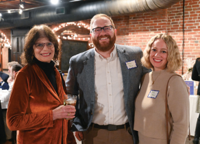 Three people standing together at an indoor event with exposed brick walls and string lights. One person is wearing a rust-colored velvet jacket and holding a drink, another is in a gray blazer over a white shirt, and the third is in a beige top with a necklace. Two visible name tags are worn on the gray blazer and beige top. 