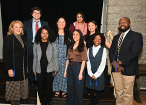 Group of nine people standing on indoor steps in front of a black curtain and stone wall. They are dressed in semi-formal and business attire, including suits, blazers, patterned dresses, and collared shirts. Several individuals have blue honor cords draped around their necks. 