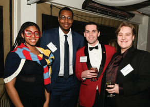 Four people standing together at an indoor event. One person is wearing a black outfit with a colorful striped scarf, another is in a dark blue suit with a white shirt and dark tie, the third is in a red tuxedo jacket with a black bow tie holding a drink, and the fourth is in a black outfit with a dark patterned scarf holding a glass. 