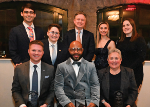 Group of eight people dressed in formal attire posing indoors in front of a stone arch and dark-framed windows. Three individuals in the front row are seated and holding glass awards, while five individuals stand behind them. Clothing includes suits, ties, and evening dresses. 