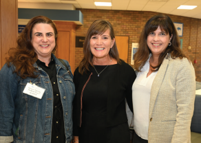 Three people standing indoors in front of a brick wall and wooden panels. One person is wearing a denim jacket over a black top with a visible name tag, another is in a black outfit with a necklace, and the third is in a light-colored blazer over a white blouse. 