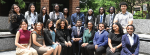 Group of people posed outdoors in front of a brick building and greenery. They are dressed in business and professional attire, seated and standing in two rows on stone benches. 