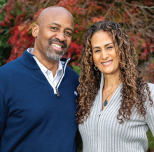 Two people standing outdoors in front of vibrant autumn foliage, wearing long-sleeve sweaters—one in navy blue and the other in light gray—with arms around each other. 