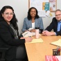 Four people sit around a rectangular wooden table in an office meeting room. The table has yellow notepads, pens, and reusable drink containers. A bulletin board with various notices and flyers is mounted on the wall behind them, and a window with blinds is on the right side of the room. 