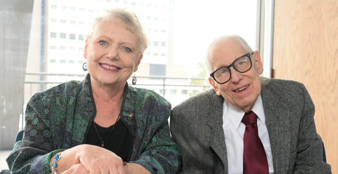Two older adults are seated side by side at a wooden table indoors. One person is wearing a patterned green jacket over a black top with jewelry on the wrist and fingers, while the other is dressed in a gray tweed jacket with a white shirt and a dark red tie. Behind them is a large window showing an urban building and a wooden door to the right. 