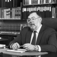 Black-and-white photo of a person seated at a desk in an office setting. The person is wearing a dark suit with a patterned tie and a wristwatch, holding a pen over a folder. The desk has several books stacked on the left side. Behind the person is a bookshelf filled with binders and books. 