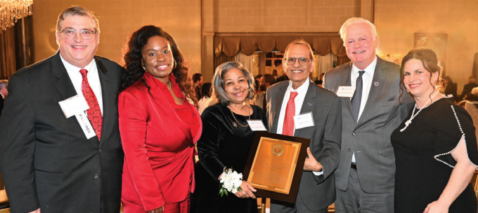 Six individuals standing together in a formal event setting with patterned carpet and chandeliers. They are dressed in formal attire, including suits and dresses, and wearing name tags. One person in the center is holding a large wooden plaque with an engraved emblem. The background shows banquet tables and warm lighting. 