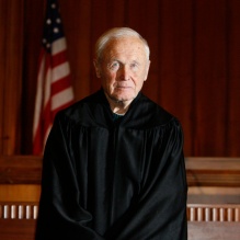 Person wearing a black judicial robe standing in a courtroom. Behind them is a wooden judge&rsquo;s bench with an American flag and a lit lamp visible in the background. The setting includes wood-paneled walls and a formal courtroom atmosphere. 