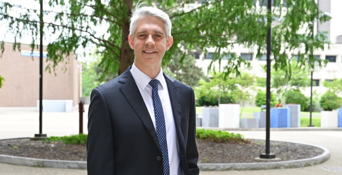 A person wearing a black business suit with a white dress shirt and a patterned tie stands outdoors on a paved walkway. The background includes landscaped areas with mulch and greenery, several trees, lamp posts, and modern campus buildings. 