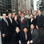A group of people dressed in formal business attire standing together on a city sidewalk in an urban setting. Tall office buildings line the street, and the Radio City Music Hall sign is visible in the background. The group appears posed for a photo, with some standing and others kneeling in front. 