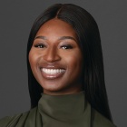 Person wearing a dark green high-neck top with long sleeves, posed against a plain dark gray background. The individual has straight, long dark hair parted in the center. 