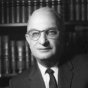 A black-and-white photograph of a person seated in front of shelves filled with books. The person is wearing a dark suit with a light-colored dress shirt and a tie, with hands resting on one knee in a formal pose. 