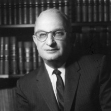 A black-and-white photograph of a person seated in front of shelves filled with books. The person is wearing a dark suit with a light-colored dress shirt and a tie, with hands resting on one knee in a formal pose. 