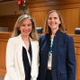 Two people standing in a courtroom setting with wooden judge&rsquo;s bench and chairs in the background. One person is wearing a white blazer over a dark dress, and the other is wearing a navy pantsuit with a patterned blouse. 