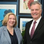 Two people in professional business attire stand indoors in front of a dark blue wall decorated with framed artwork. One person is wearing a light blue textured blazer over a dark top with a pearl necklace, and the other is wearing a dark pinstripe suit with a white shirt and maroon patterned tie. 