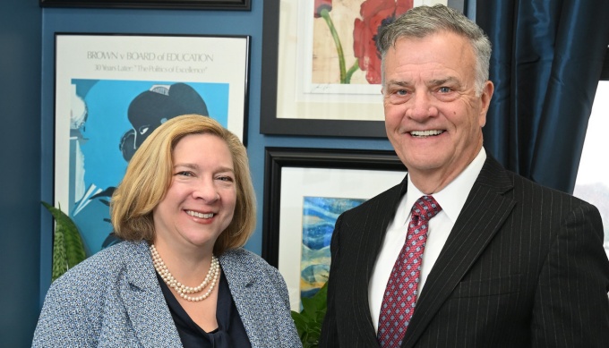 Two people in professional business attire stand indoors in front of a dark blue wall decorated with framed artwork. One person is wearing a light blue textured blazer over a dark top with a pearl necklace, and the other is wearing a dark pinstripe suit with a white shirt and maroon patterned tie. There are potted plants visible near the floor. 