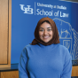 Person wearing a brown headscarf and a bright blue long-sleeve sweater with a black belt, standing indoors in front of a blue backdrop with the University at Buffalo School of Law logo and wooden panels. 