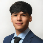 Studio portrait of a person wearing a blue suit jacket, light blue dress shirt, and black tie against a light background. 